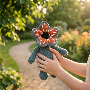 May include: A handmade crochet Demogorgon doll. The doll features a green body and arms, with a distinctive flower-like head in shades of orange, white, and green. The doll is held in front of a blurred outdoor background.