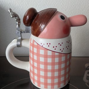 Peut inclure: Une chope à bière en céramique fantaisie avec un design de visage de dessin animé. La chope présente un motif à carreaux roses et blancs, une anse et un couvercle à charnière. Le visage a un long nez, des cheveux bruns et un teint rose.