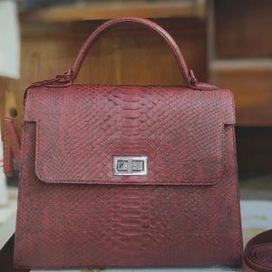 May include: A burgundy leather handbag with a snakeskin pattern. The bag has a top handle, a flap closure with a silver clasp, and a matching shoulder strap. The bag is approximately 30 cm wide and 25 cm tall.