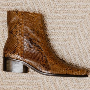 May include: A brown ankle boot with a snakeskin pattern. The boot has a square toe and a low, block heel. The boot is made of a textured material and is set against a neutral background.