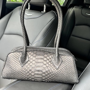 Puede incluir: Un bolso de mano gris carbón metalizado con un patrón texturizado de piel de serpiente en todo el exterior. El bolso tiene un cuerpo rectangular con cierre de cremallera superior y dos asas trenzadas a juego unidas al panel superior.