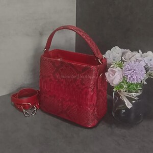 May include: A red leather handbag with a top handle and a detachable shoulder strap. The bag has a textured snakeskin pattern and silver-tone hardware. The text "Djakarta Boutique" is visible on the side of the bag. A small bouquet of flowers is in the background.