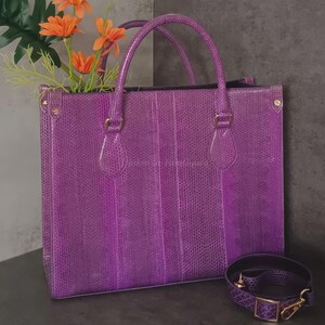 May include: A vibrant purple tote bag with matching shoulder strap. The bag features a textured, reptile-like pattern, double handles, and gold-colored hardware. Orange flowers peek out from the top of the bag, adding a pop of color.