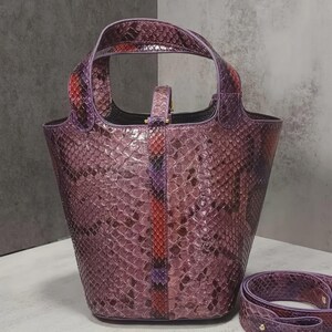 May include: A purple handbag with a snakeskin pattern. The bag has a structured bucket shape with a top handle and a detachable shoulder strap. The strap has a gold-colored buckle. The bag's color is a mix of purple, red, and orange.