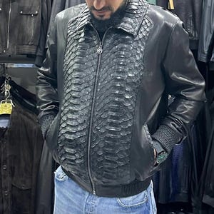 Könnte beinhalten: Schwarze Lederjacke mit Schlangenleder-Struktur auf der Vorderseite und am Kragen. Die Jacke hat einen Reißverschluss, gerippte Bündchen und einen gerippten Saum. Die Ärmel und der Rücken sind aus glattem schwarzem Leder. Die Jacke wird in einem Bekleidungsgeschäft ausgestellt.