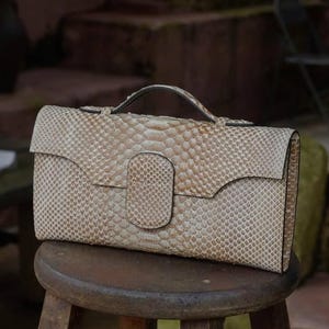 Puede incluir: Un bolso de mano beige de piel de serpiente con asa y cierre de solapa. El bolso es rectangular y está sobre un taburete de madera. El patrón de piel de serpiente es visible en la superficie del bolso.
