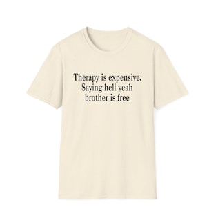 以下が含まれることがあります： わずかに斑点のある質感のクリーム色の半袖Tシャツ。前面には「Therapy is expensive. Saying hell yeah brother is free.」という黒いテキストがプリントされています。Tシャツはクルーネックで、標準的なストレートカットのシルエットです。