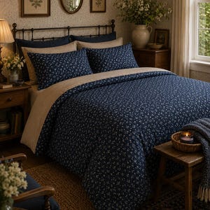 Op de afbeelding: Een bed met een marineblauw dekbedovertrek en bijpassende kussenslopen met een klein wit bloemenpatroon. Het beddengoed is gelaagd met effen beige lakens en kussenslopen, geplaatst tegen een donker metalen hoofdeinde in een slaapkamer.