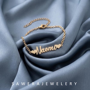 May include: A gold-toned metal bracelet featuring a delicate chain and a central nameplate with the name Naomi in cursive script, flanked by two small heart shapes. The bracelet includes a lobster claw clasp and an adjustable extender chain.