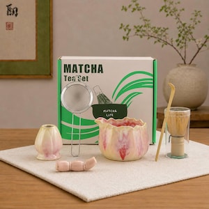 Puede incluir: Juego de té Matcha con una caja que dice "Matcha Tea Set". El juego incluye un cuenco de cerámica, un batidor, un colador y otros accesorios. El cuenco es rosa y amarillo con un borde con volantes. El juego está sobre un paño blanco.