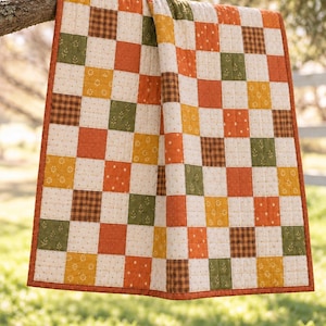 Peut inclure: Une courtepointe en patchwork composée d'une grille de carrés de tissu aux motifs et couleurs alternés, incluant de l'orange uni, du carreau marron, du crème à petites fleurs et du vert olive à motifs de feuilles, avec une bordure orange.