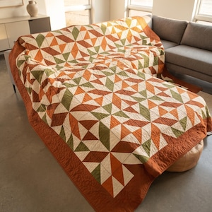 May include: A quilt featuring a geometric star pattern composed of triangular fabric patches in shades of burnt orange, olive green, and cream. The textile has a solid burnt orange border and visible quilted stitching throughout the entire surface.