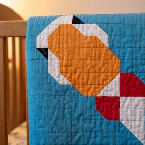 May include: A quilted blanket featuring a vibrant blue background with a geometric pattern composed of orange, white, and red fabric squares. The quilt displays a grid-like stitching texture throughout the surface, highlighting the pieced construction.