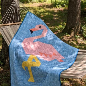 Op de afbeelding: Een doorgestikte deken met een geometrisch roze flamingo-ontwerp op een gemêleerde lichtblauwe achtergrond. De flamingo heeft een zwart knoopoog, een zwart-oranje snavel en een geel been. De quilt heeft een rasterachtig stikpatroon.