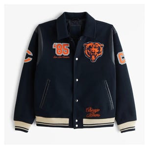 Op de afbeelding: Marineblauwe varsity jas met oranje en witte accenten. Voorzien van het nummer '85' en een berenlogo op de voorkant, met het opschrift 'Chicago Bears'. Inclusief leren mouwen en een knoopsluiting.