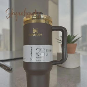 Puede incluir: Un vaso Stanley Quencher H2.0 marr&oacute;n y dorado con asa. El vaso tiene una tapa dorada y una etiqueta blanca con el texto "QUENCHER H2.0 TUMBLER" y "1,18L". El logotipo de Stanley es visible.