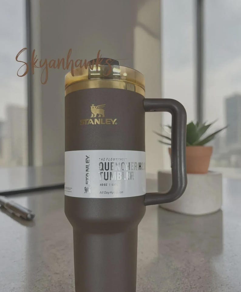 Puede incluir: Un vaso Stanley Quencher H2.0 marr&oacute;n y dorado con asa. El vaso tiene una tapa dorada y una etiqueta blanca con el texto "QUENCHER H2.0 TUMBLER" y "1,18L". El logotipo de Stanley es visible.