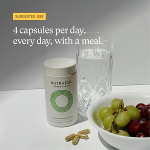 May include: A white cylindrical bottle of Nutrafol Women's Vegan hair growth supplement with green circular design, next to a glass of water, a bowl of grapes, and four capsules. The text on the bottle reads "4 capsules per day, every day, with a meal."