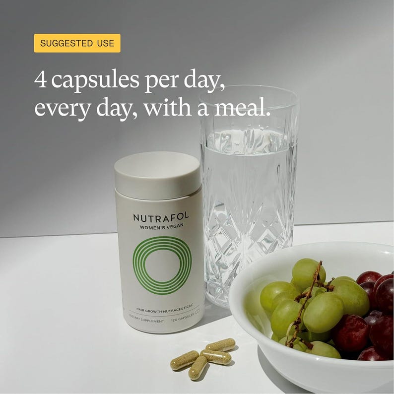 May include: A white cylindrical bottle of Nutrafol Women's Vegan hair growth supplement with green circular design, next to a glass of water, a bowl of grapes, and four capsules. The text on the bottle reads "4 capsules per day, every day, with a meal."