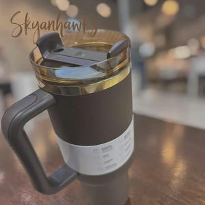 Puede incluir: Un vaso t&eacute;rmico negro con tapa transparente y una banda dorada. El vaso tiene un asa negra y una banda blanca en el centro. El texto "Skyanhawks" est&aacute; escrito en la parte superior izquierda.