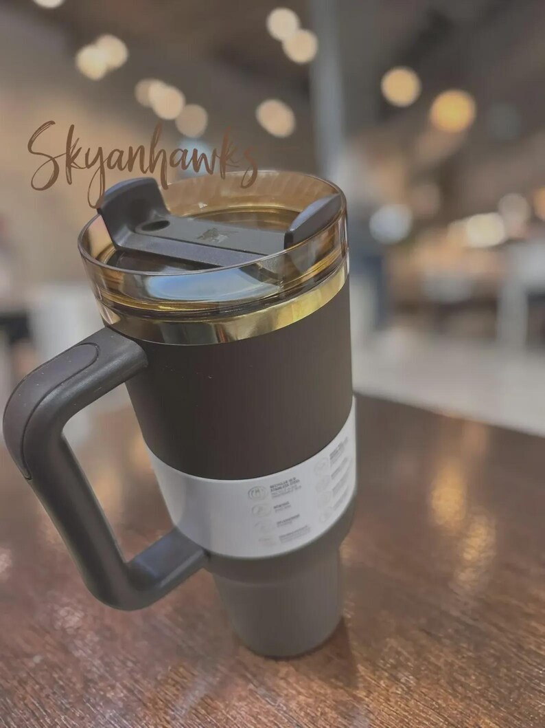 Puede incluir: Un vaso t&eacute;rmico negro con tapa transparente y una banda dorada. El vaso tiene un asa negra y una banda blanca en el centro. El texto "Skyanhawks" est&aacute; escrito en la parte superior izquierda.