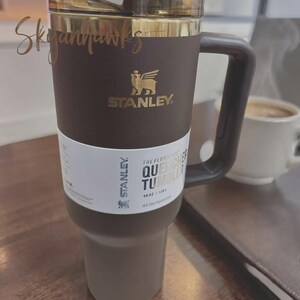 Puede incluir: Un vaso Stanley Quencher marr&oacute;n con tapa y asa doradas. El vaso tiene el logotipo de Stanley y las palabras "THE FLOWSTATE QUENCHER TUMBLER" impresas. Est&aacute; dise&ntilde;ado para la hidrataci&oacute;n durante todo el d&iacute;a y tiene una capacidad de 1,18L.