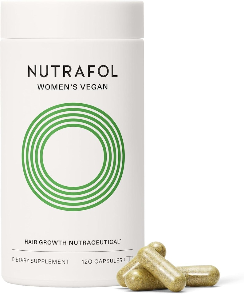 May include: A white cylindrical bottle of Nutrafol Women's Vegan hair growth nutraceutical dietary supplement. The bottle has a green circular design and the text "120 Capsules". Three capsules are in front of the bottle.