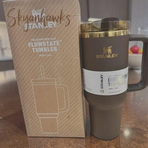 Puede incluir: Un vaso Stanley Flowstate marr&oacute;n con asa y tapa dorada, junto a su caja de cart&oacute;n. La caja presenta el texto "Skyanhawks" y un dibujo del vaso. El vaso contiene 1,18L.