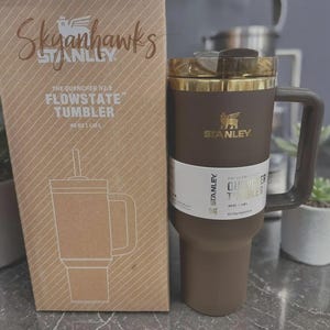 Puede incluir: Un vaso Stanley Flowstate marr&oacute;n con asa y tapa dorada, junto a una caja de cart&oacute;n con el texto "Skyhawks" y una ilustraci&oacute;n del vaso. El vaso tiene una capacidad de 1,18L.