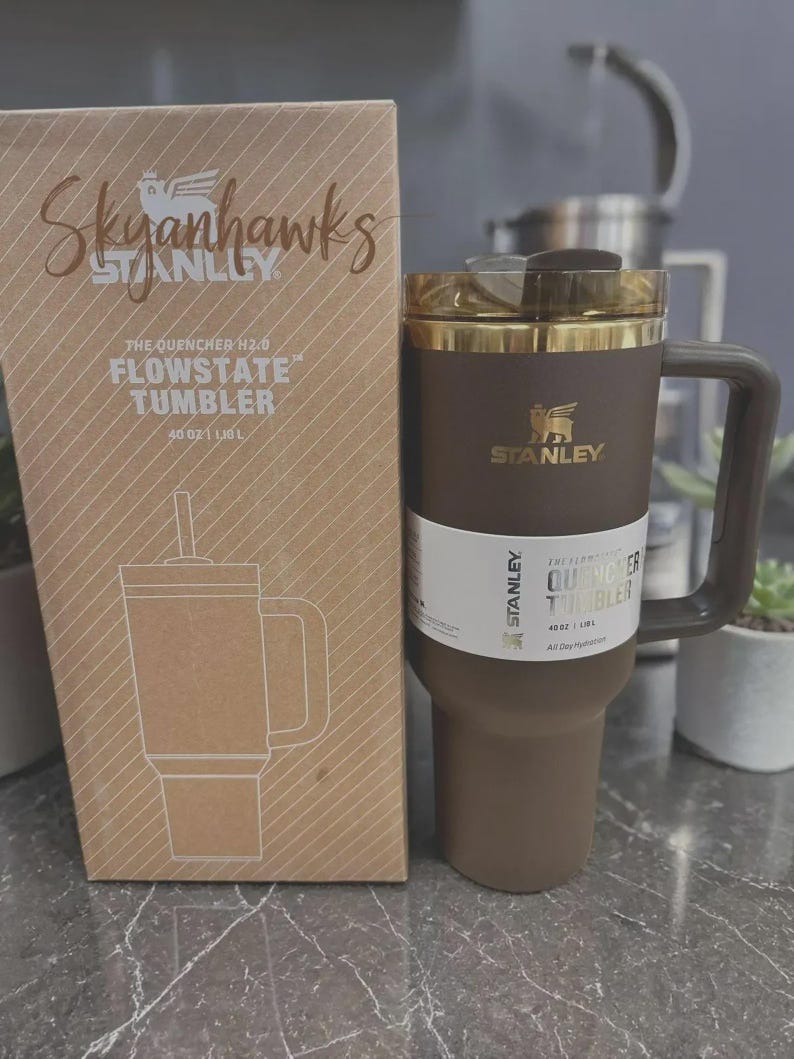 Puede incluir: Un vaso Stanley Flowstate marr&oacute;n con asa y tapa dorada, junto a una caja de cart&oacute;n con el texto "Skyhawks" y una ilustraci&oacute;n del vaso. El vaso tiene una capacidad de 1,18L.