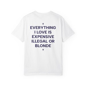 Può includere: Una t-shirt bianca a maniche corte con testo blu scuro sul retro che recita EVERYTHING I LOVE IS EXPENSIVE ILLEGAL OR BLONDE, centrato tra due piccoli simboli a stella a quattro punte disposti verticalmente sopra e sotto il testo.