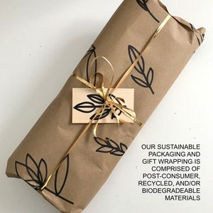 May include: A brown paper gift package with a gold ribbon and a small tag. The paper has a black and white leaf pattern. The text on the tag reads: "OUR SUSTAINABLE PACKAGING AND GIFT WRAPPING IS COMPRISED OF POST-CONSUMER, RECYCLED, AND/OR BIODEGRADEABLE MATERIALS."