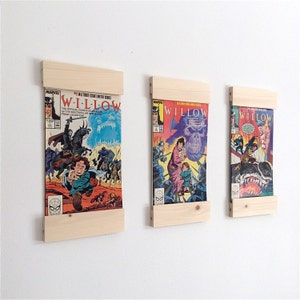 May include: Three wooden comic book display shelves with three comic books featuring the title "WILLOW" and images of characters from the film. The comic books are displayed on the shelves with the covers facing out.