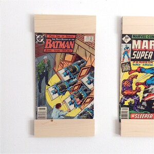 May include: Two comic books are displayed on wooden shelves. The left comic book is a DC Comics Batman issue with the title "The Many Deaths of the Batman". The right comic book is a Marvel Comics issue featuring Captain America and the title "The Sleeper".
