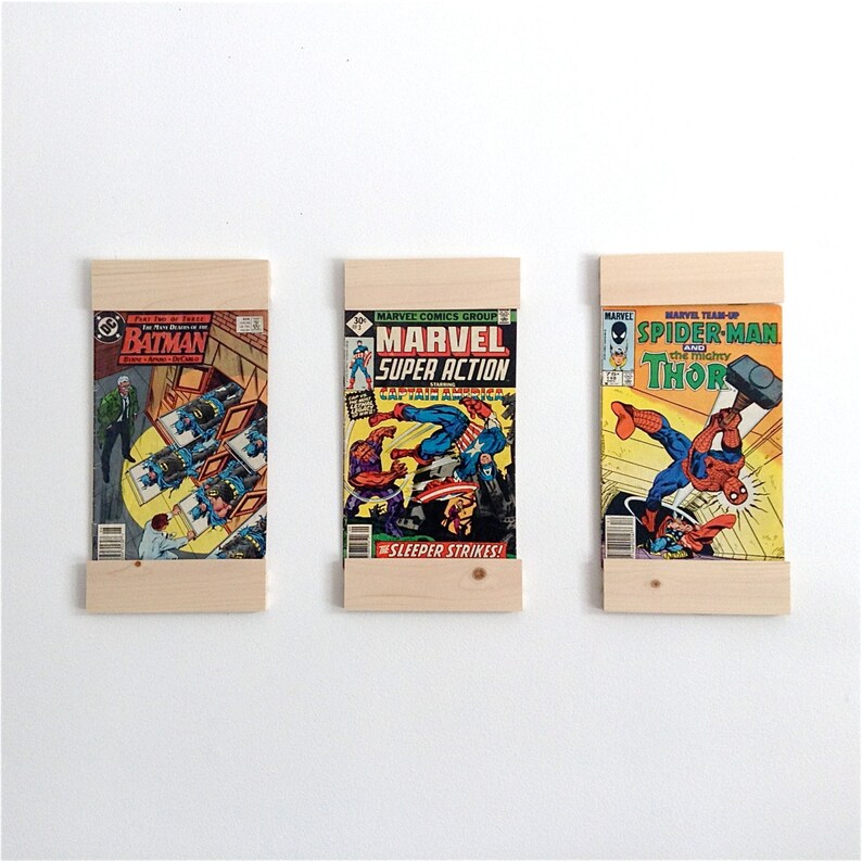 May include: Three comic books are displayed on a wall using a wooden shelf system. The comic books are "Batman", "Marvel Super Action", and "Spider-Man and the mighty Thor".