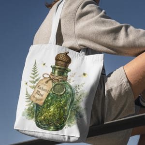 May include: A white tote bag featuring a graphic illustration of a green glass bottle filled with moss and glowing lights. The bottle has an acorn-shaped stopper, a twine-tied tag labeled Enchanted Moss, and is surrounded by fern leaves and bees.