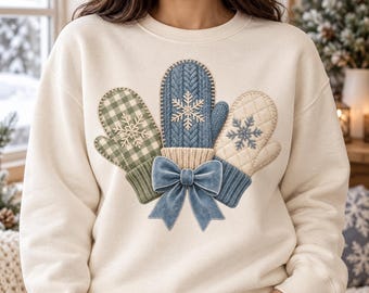 May include: A cream-colored crewneck sweatshirt featuring a central graphic of three mittens tied together with a blue velvet bow. The mittens display distinct patterns: green gingham, blue cable knit, and quilted cream, each with a snowflake.