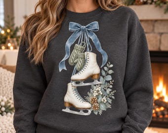 May include: A dark charcoal gray crewneck sweatshirt featuring a graphic print of two white ice skates hanging from a large light blue ribbon bow. The design includes a pair of sage green knit mittens, pinecones, and winter foliage.