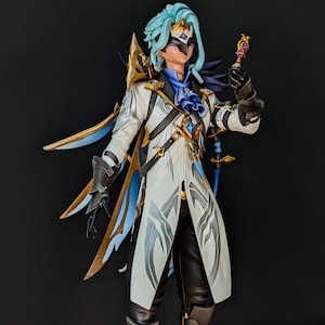 Puede incluir: Una figura coleccionable de un personaje con cabello azul claro, vistiendo un abrigo blanco con detalles dorados y azules, una máscara oscura y guantes negros. La figura sostiene un pequeño cetro adornado en violeta y oro y presenta intrincadas placas de armadura en los hombros y la espalda.