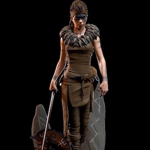 May include: A brown-clad warrior figurine featuring a feathered collar, a headpiece with a central emblem, and dark eye makeup. The figure holds a long sword and stands beside a circular shield with etched markings and two large stone-like slabs.