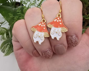 May include: A pair of gold-colored mushroom earrings. Each earring features a white mushroom body with a smiling face and a red cap with white spots. The earrings are held up by gold-colored hooks.