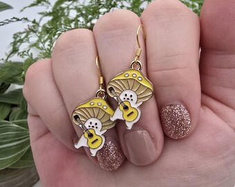 May include: A pair of gold-colored mushroom earrings. Each earring features a yellow mushroom cap with white spots and a white figure playing a yellow guitar. The earrings are held up by gold-colored hooks.