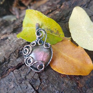 May include: A silver wire wrapped pendant featuring a dark grey and pink labradorite gemstone. The pendant is resting on a piece of bark with autumn leaves.