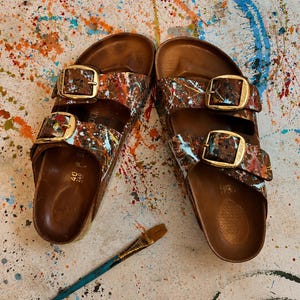May include: A pair of brown leather sandals with a colorful paint splatter design. The sandals have two adjustable straps with gold-colored buckles. A small paintbrush with a blue handle and gold ferrule rests nearby.
