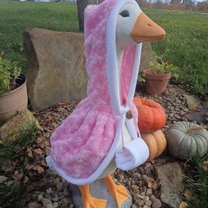 May include: A white goose statue wearing a pink, textured, hooded cape with a white trim. The cape features a small, circular, metallic-colored button closure at the neck and a white muff-style accessory hanging from the front of the garment.