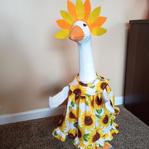 Könnte beinhalten: Eine weiße Gänsefigur mit einem Kleid mit Sonnenblumenmuster und einem Sonnenblumenkranz. Das Kleid hat einen weißen Hintergrund mit großen gelben Sonnenblumen. Die Gans hat einen orangefarbenen Schnabel und Füße. Der Kranz ist gelb und orange.