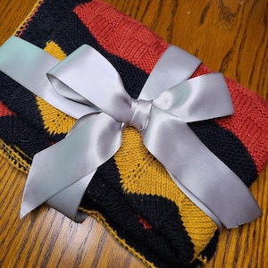 May include: A folded knitted blanket featuring a geometric pattern in black, mustard yellow, and burnt orange, tied with a wide, smooth, light gray satin ribbon that is fashioned into a large decorative bow across the center of the textile.