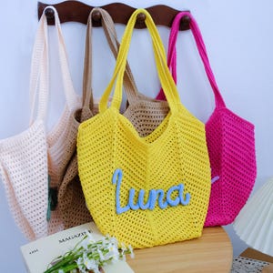 May include: Four crochet tote bags in light pink, beige, bright yellow, and hot pink hanging on a wooden rack. The yellow bag features the name Luna in light blue cursive yarn lettering stitched onto the front. Each bag has a textured open-knit pattern.