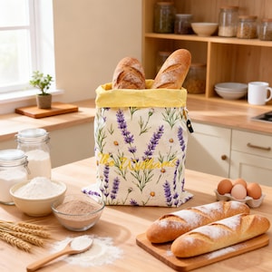 Puede incluir: Una bolsa de tela color crema para pan con un estampado floral de lavanda y margaritas y un forro interior amarillo. La bolsa está bordada con el texto "The Williams" en letra cursiva amarilla y contiene dos barras de pan.