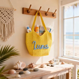 May include: A bright yellow crochet tote bag featuring the name Luna in light blue cursive lettering on the front. The bag contains a white bottle, an orange bottle, and a book, and is hanging from a wooden hook on a light-colored wall.